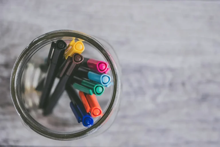 Top view of colourful pens in a glass jar on a wooden surface.