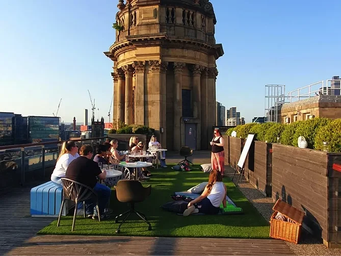 A group of people attends a psychotherapy workshop on a rooftop terrace in the late afternoon sun. Participants are seated on chairs around tables or on picnic blankets spread over artificial grass. A facilitator stands near a flip chart, speaking to the group. The backdrop features a large historic stone tower with classical architectural details, contrasting with the modern city skyline in the distance. The setting feels relaxed and open, encouraging reflection and conversation.