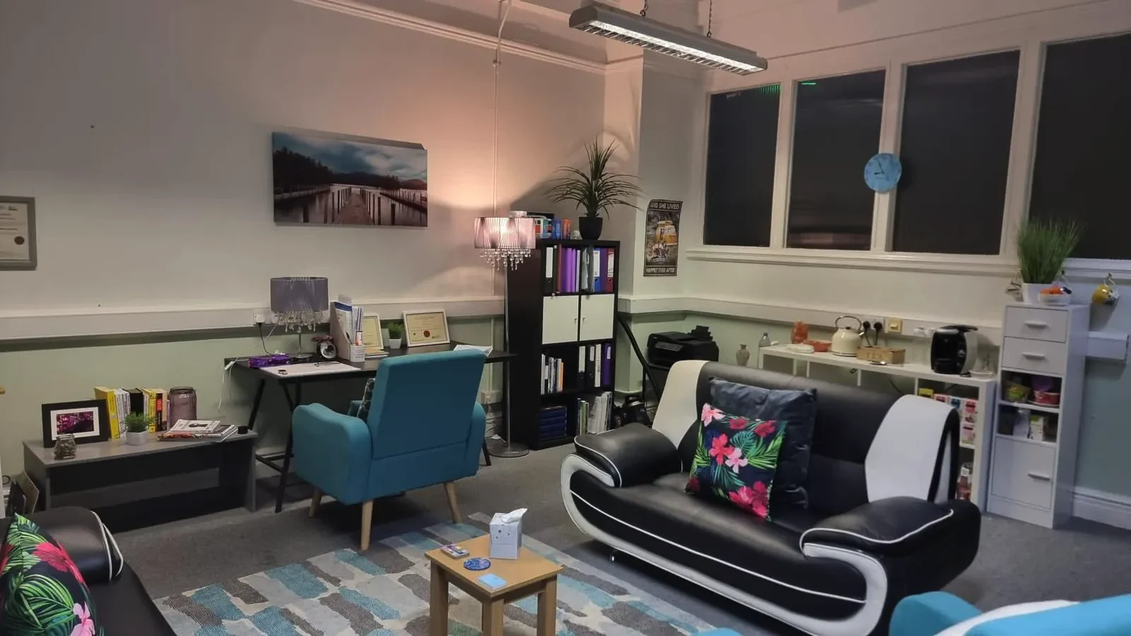 Therapy office with a sofa, armchairs, desk, bookshelf, and tea station. Calm lighting and decorative touches throughout.